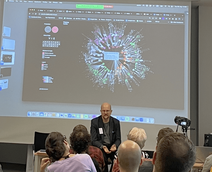 A man sits in front of a large screen providing a lecture. The screen has a projection of a network map.