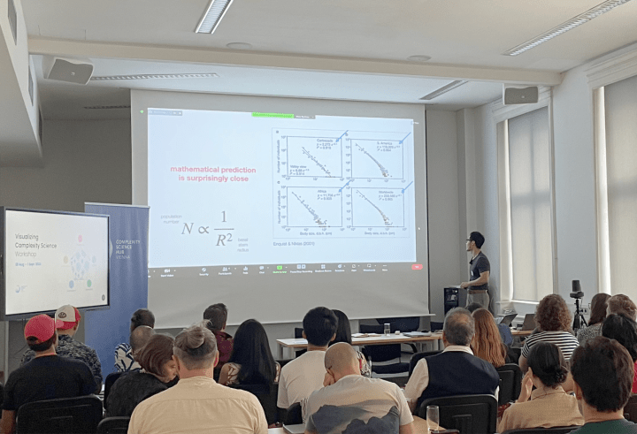 A man sits in front of a large screen providing a lecture. The screen has a projection of a scatter plots with the text 